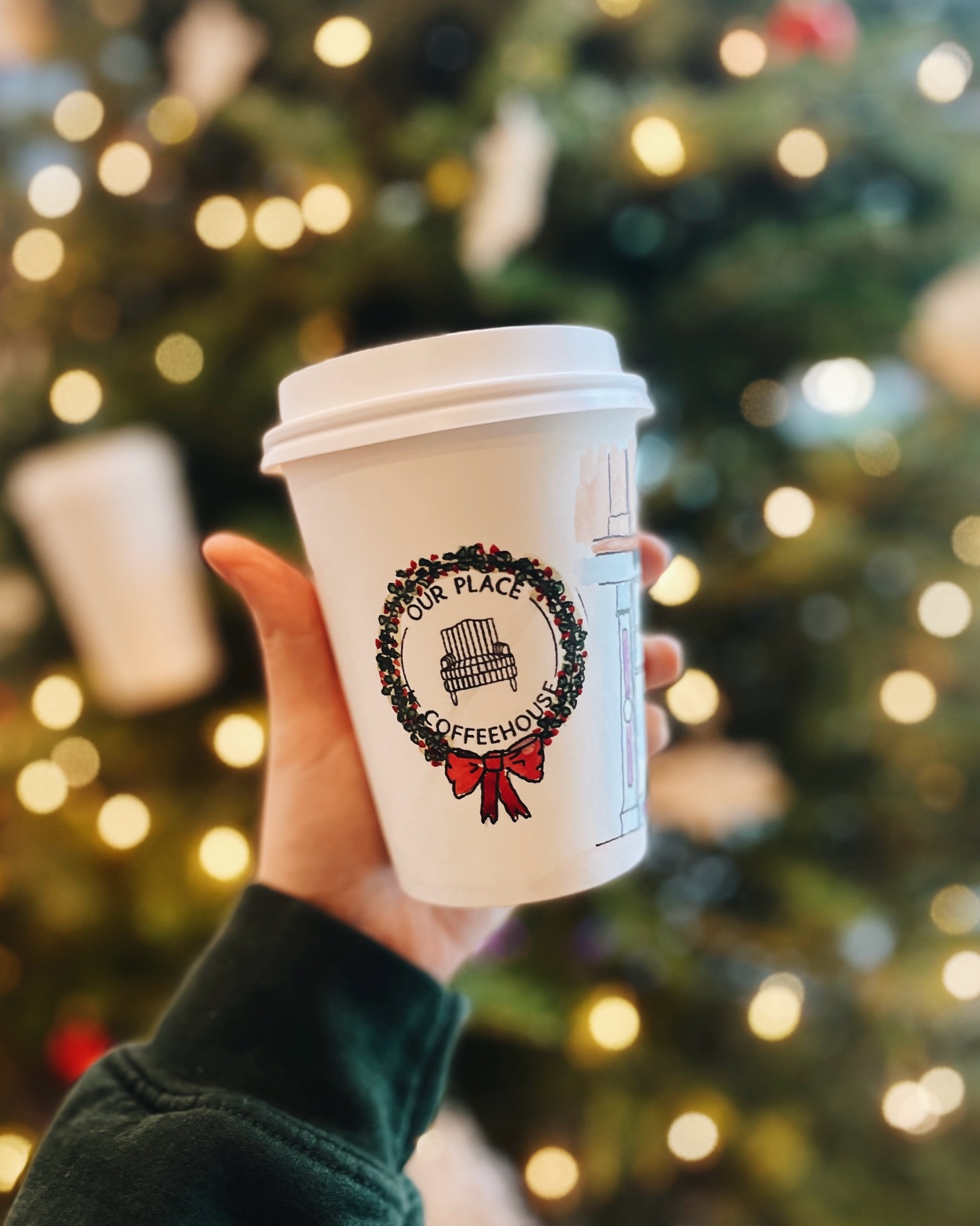An Our Place cup painted with a holiday wreath in front of warm Christmas lights